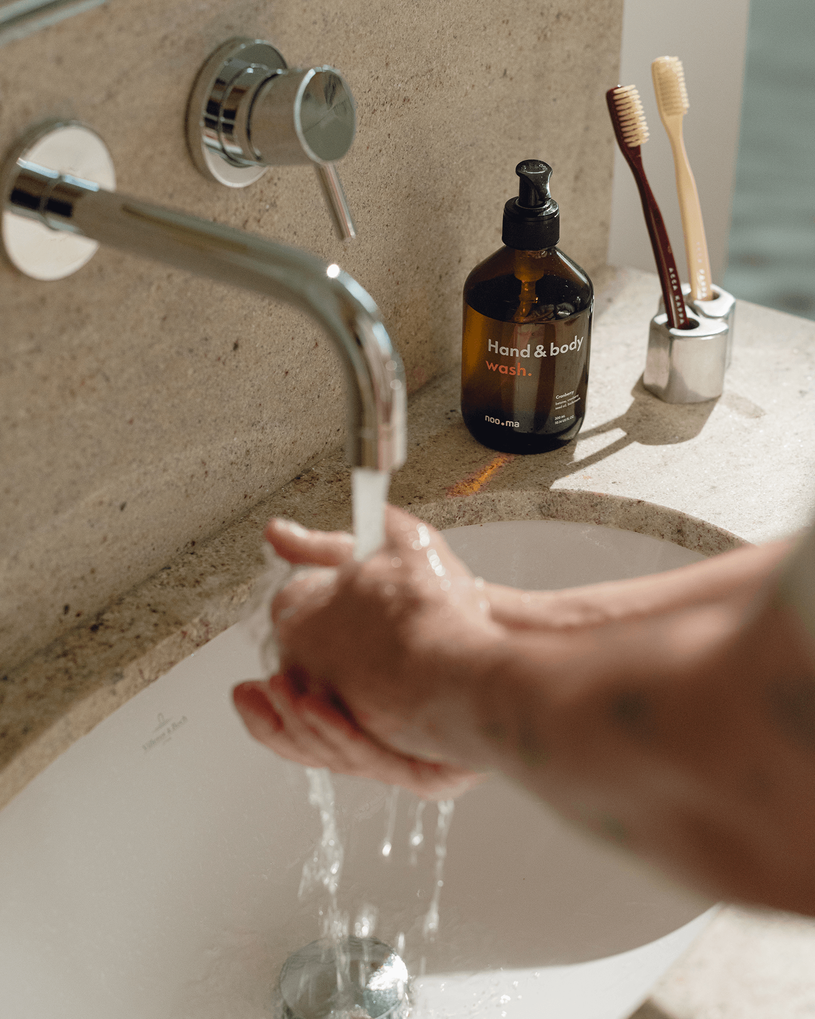 Person washing hands under running water in a sink with a bottle of noo.ma's Hand & Body Soap bottle and toothbrushes in the background.